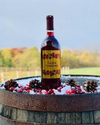 A bottle of Frosted Cranberry Wine sits on a wine barrel with snow and pinecones.