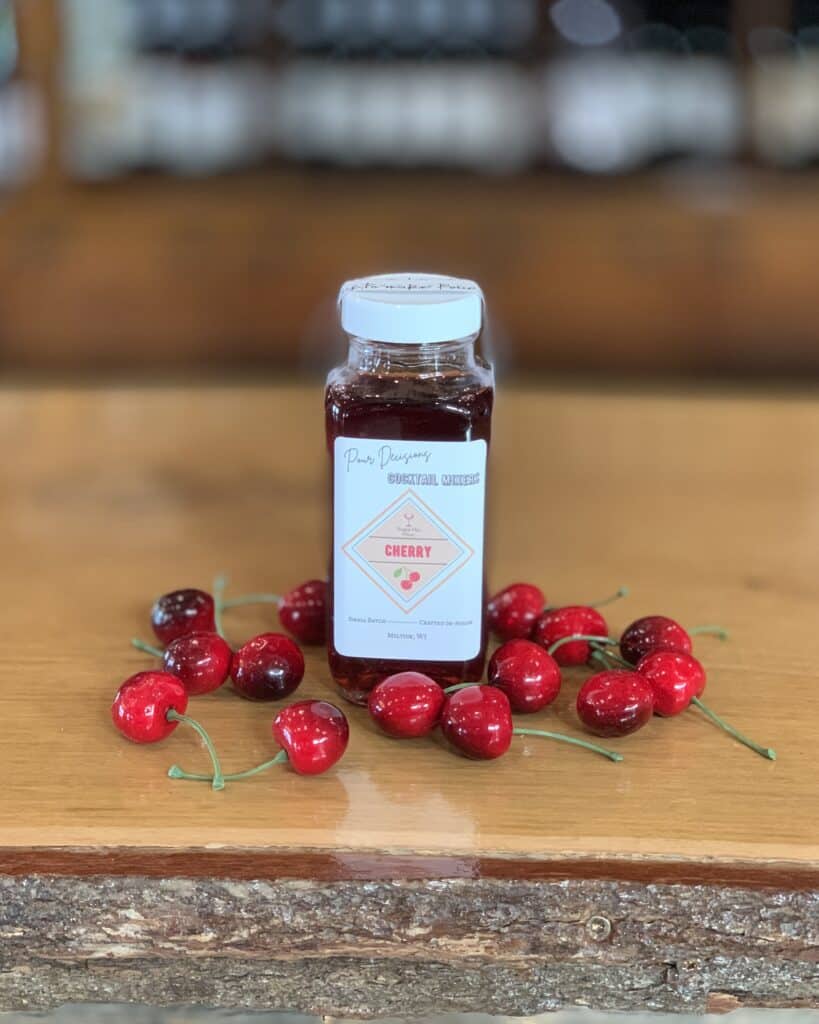 A bottle of cherry cocktail mixer surrounded by ripe, red cherries.