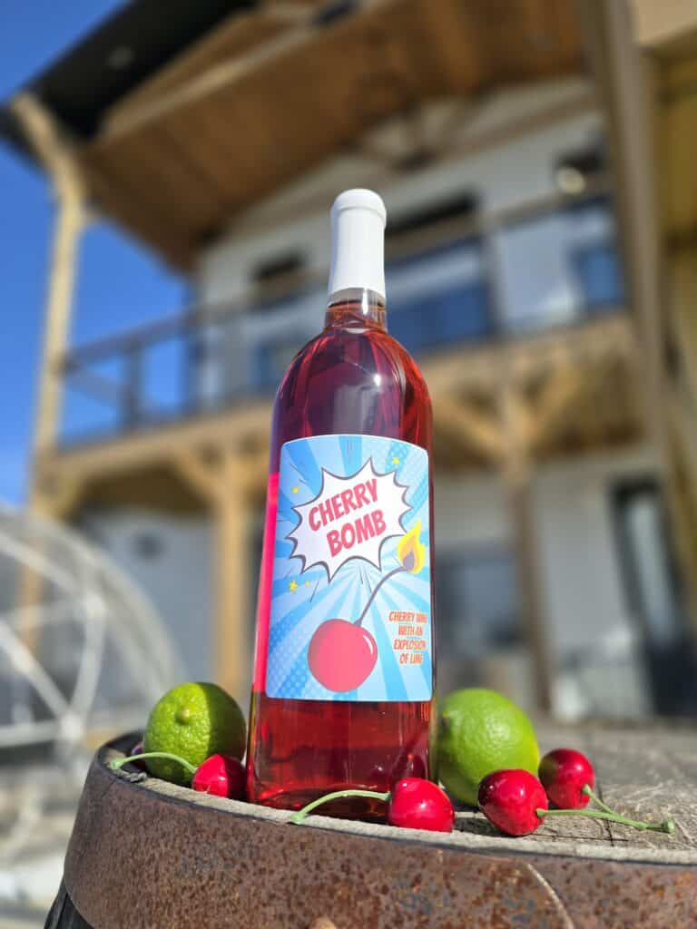 A bottle of bright red Cherry Bomb Wine sits on a barrel amongst some limes and cherries. There is a bright blue sky in the background framing the beautiful Timber Hill Winery building.