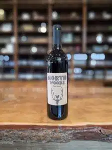 A bottle of Northwoods Red wine sits on a wooden countertop with shelves of wine bottles in the background.